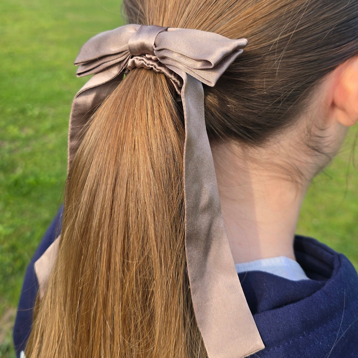 Person with long hair tied back with a ribbon, outdoors with greenery in the background